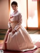 A woman in a white and pink hanbok sitting on a red mat.